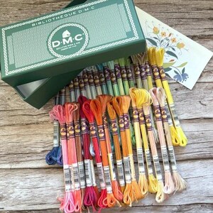 May include: A collection of colorful DMC embroidery floss skeins in various shades of pink, orange, yellow, and blue, arranged near an open green box with the text "BIBLIOTHEQUE D.M.C." and a floral card.