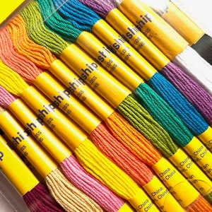 May include: A close-up of a rainbow assortment of embroidery floss spools. The spools are labeled "FLOSS" and "STRANDS" and are packaged in a clear plastic container. The spools are arranged in a rainbow pattern, with the colors red, orange, yellow, green, blue, indigo, and violet.