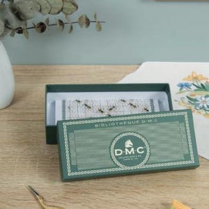 Könnte beinhalten: Eine grüne Schachtel mit der Aufschrift "BIBLIOTHEQUE D.M.C" und einem D.M.C-Logo, die ein Kreuzstichprojekt mit Bienen enthält. Goldene Scheren und Stickgarn liegen auf einer Holzoberfläche. Eine Blumenstickerei befindet sich im Hintergrund.