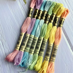May include: A set of eight embroidery floss spools in pastel colors, including pink, lavender, light blue, green, yellow, and orange. Each spool has a white label with black text that includes the brand name "DMC" and the number of the floss color.