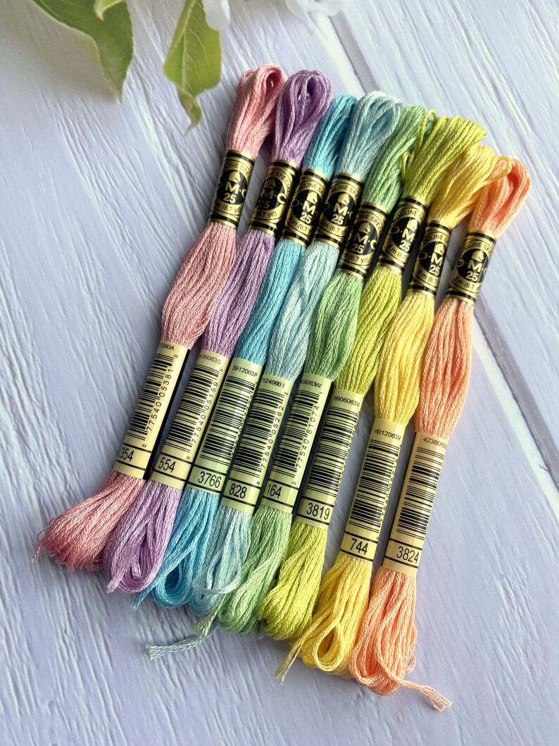 May include: A set of eight embroidery floss spools in pastel colors, including pink, lavender, light blue, green, yellow, and orange. Each spool has a white label with black text that includes the brand name "DMC" and the number of the floss color.