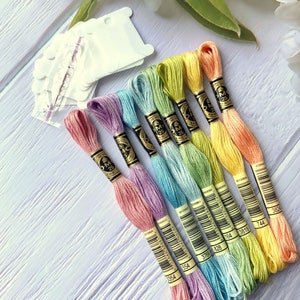 May include: A set of eight embroidery floss spools in a rainbow of pastel colors, including pink, lavender, light blue, turquoise, green, yellow, and orange. Each spool has a black and gold label with the DMC brand name and a barcode. The spools are arranged in a fan shape on a white wooden surface.