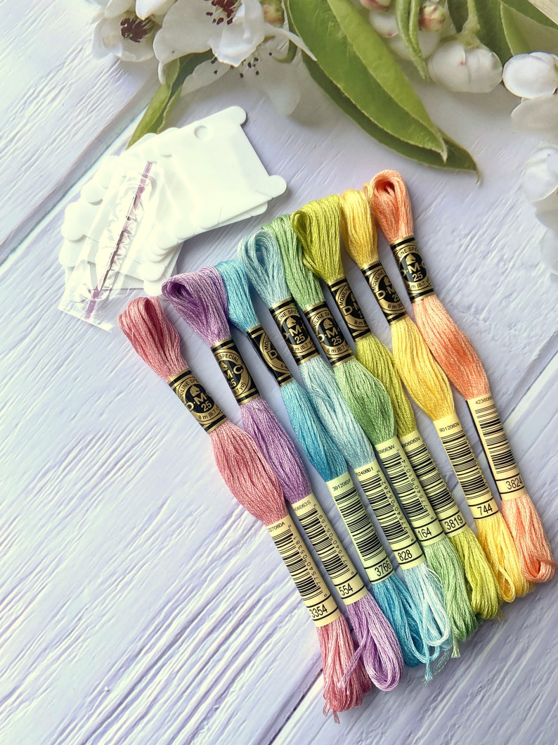 May include: A set of eight embroidery floss spools in a rainbow of pastel colors, including pink, lavender, light blue, turquoise, green, yellow, and orange. Each spool has a black and gold label with the DMC brand name and a barcode. The spools are arranged in a fan shape on a white wooden surface.