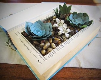 May include: A book with a blue cover has been repurposed into a decorative planter. The book is open and the pages have been cut out to create a small garden. The garden is filled with small stones, paper flowers, and two blue paper succulents.
