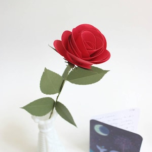 May include: A single red paper rose in a white milk glass vase. The rose is made of multiple layers of paper, creating a realistic look. The vase is a classic milk glass design with a textured surface.