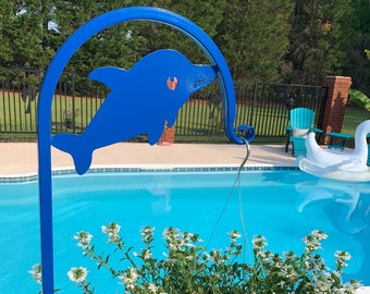 Könnte beinhalten: Blauer, metallener, delphin-förmiger Hängepflanzenständer mit einer weißen blühenden Pflanze, die daran hängt. Der Ständer steht vor einem Schwimmbad.