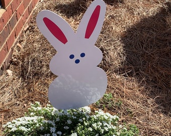 Könnte beinhalten: Eine weiße Osterhasen-Gartendekoration mit rosa Innenohren und blauen Augen. Der Hase steht vor einem Busch mit kleinen weißen Blüten, umgeben von braunen Kiefernnadeln.