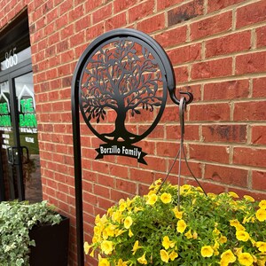 May include: Black metal tree of life design with a hanging planter. The planter is filled with yellow flowers. The tree of life has a banner that reads "Borzillo Family".