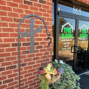 May include: A black metal plant stand with a cross design and a hanging planter filled with succulents and green plants. The stand is in front of a brick building with a sign that reads "The Barn of All Trades, LLC".