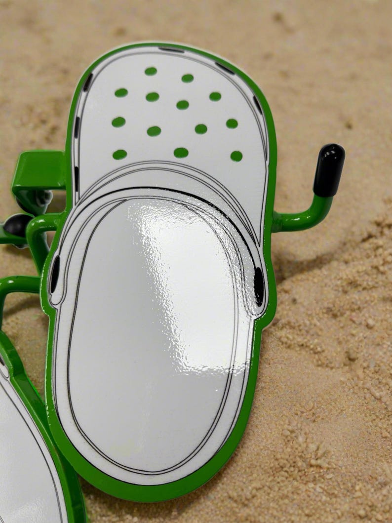 K&ouml;nnte beinhalten: Ein gr&uuml;ner Wandhaken aus Metall in Form eines wei&szlig;en Clogs mit gr&uuml;nen Punkten. Der Haken hat eine gl&auml;nzende Oberfl&auml;che, einen seitlich abstehenden Arm mit schwarzer Spitze und ein flaches, stilisiertes Design, das einem Clog nachempfunden ist.