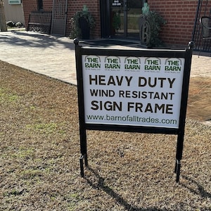 May include: A black sign frame with the text "Heavy Duty Wind Resistant Sign Frame" and the website address "www.barnofalltrades.com". The sign is in front of a brick building with a green plant in a pot.