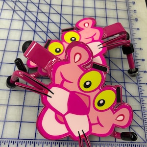 K&ouml;nnte beinhalten: Zwei Wandhaken aus Metall in Form der Figur des Pink Panther. Jeder Haken zeigt ein rosa Gesicht mit gelben Augen, schwarzen Augenbrauen und schwarzen Schnurrhaaren. Die Haken haben eine gl&auml;nzende Oberfl&auml;che und schwarze Gummikappen an den Enden.