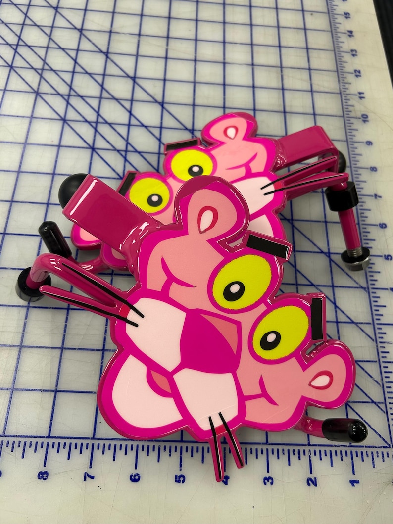 K&ouml;nnte beinhalten: Zwei Wandhaken aus Metall in Form der Figur des Pink Panther. Jeder Haken zeigt ein rosa Gesicht mit gelben Augen, schwarzen Augenbrauen und schwarzen Schnurrhaaren. Die Haken haben eine gl&auml;nzende Oberfl&auml;che und schwarze Gummikappen an den Enden.