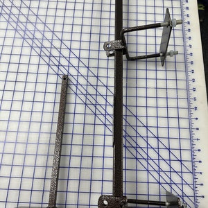 May include: A brown textured metal T-shaped stand and a long metal bar with two attached U-bolt mounting brackets. The components are positioned on a white cutting mat with blue grid lines and imperial measurements visible along the edges.