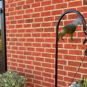 May include: Black metal silhouette of a German Shepherd dog hanging from a black metal hook. The hook is attached to a black metal stand. The stand is in front of a brick wall. The sign "The Barn of All Trades, LLC" is visible on a door behind the stand.