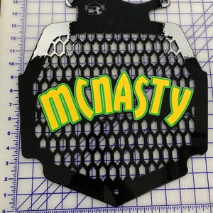 K&ouml;nnte beinhalten: Schwarzer Motorrad-K&uuml;hlergrill aus Metall mit Wabenmuster und dem Wort "MCNASTY" in gr&uuml;nen und gelben Buchstaben.