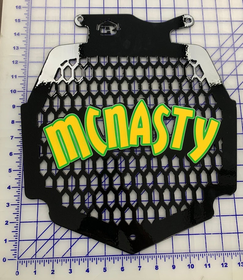 K&ouml;nnte beinhalten: Schwarzer Motorrad-K&uuml;hlergrill aus Metall mit Wabenmuster und dem Wort "MCNASTY" in gr&uuml;nen und gelben Buchstaben.