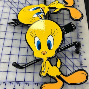 K&ouml;nnte beinhalten: Zwei gelbe Tweety Bird Anh&auml;ngerkupplungsabdeckungen aus Metall mit schwarzen Umrandungen und blauen Augen. Jede Figur hat eine gl&auml;nzende Oberfl&auml;che und ist an einer schwarzen Metallhalterung befestigt. Die Objekte liegen auf einer wei&szlig;en Schneidematte.