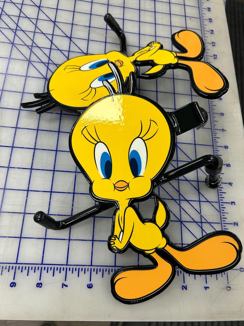 K&ouml;nnte beinhalten: Zwei gelbe Tweety Bird Anh&auml;ngerkupplungsabdeckungen aus Metall mit schwarzen Umrandungen und blauen Augen. Jede Figur hat eine gl&auml;nzende Oberfl&auml;che und ist an einer schwarzen Metallhalterung befestigt. Die Objekte liegen auf einer wei&szlig;en Schneidematte.