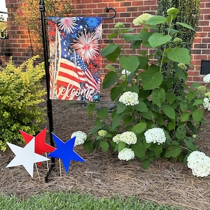 May include: A rectangular garden flag featuring a graphic of the American flag and red, white, and blue fireworks with the word welcome in white script. The flag hangs on a black metal stand next to three star-shaped yard stakes in red, white, and blue.