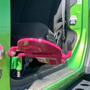 K&ouml;nnte beinhalten: Eine leuchtend pinke Fu&szlig;raste aus Metall, die am unteren T&uuml;rscharnier eines lindgr&uuml;nen Fahrzeugs befestigt ist. Die Fu&szlig;raste verf&uuml;gt &uuml;ber eine geschwungene, flache Auflagefl&auml;che f&uuml;r den Fu&szlig; und einen kleinen Haken mit schwarzer Gummikappe am Ende.