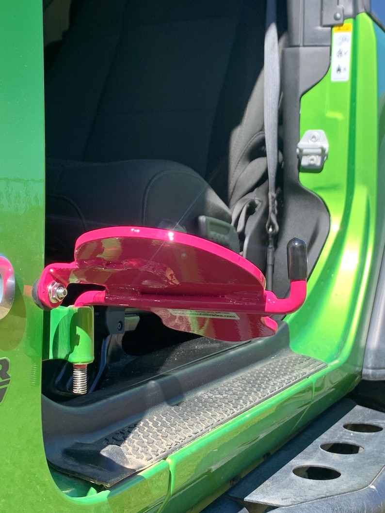 K&ouml;nnte beinhalten: Eine leuchtend pinke Fu&szlig;raste aus Metall, die am unteren T&uuml;rscharnier eines lindgr&uuml;nen Fahrzeugs befestigt ist. Die Fu&szlig;raste verf&uuml;gt &uuml;ber eine geschwungene, flache Auflagefl&auml;che f&uuml;r den Fu&szlig; und einen kleinen Haken mit schwarzer Gummikappe am Ende.