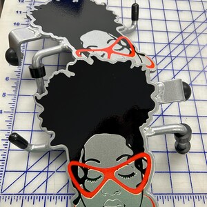 K&ouml;nnte beinhalten: Zwei T&uuml;rhaken aus Metall mit einem stilisierten Gesicht mit schwarzem Haar, grauem Teint, leuchtend orangefarbener Katzenaugenbrille und passenden orangefarbenen Creolen. Die Haken sind etwa 15 cm hoch und 10 cm breit.