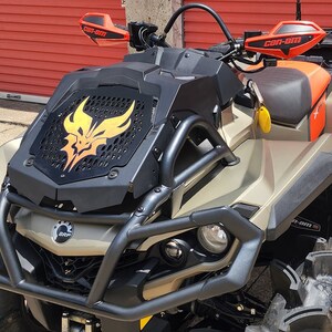 May include: A tan and black Can-Am off-road vehicle with a black and yellow graphic on the front grill. The vehicle has a roll cage and a black seat. The vehicle is on a black surface.