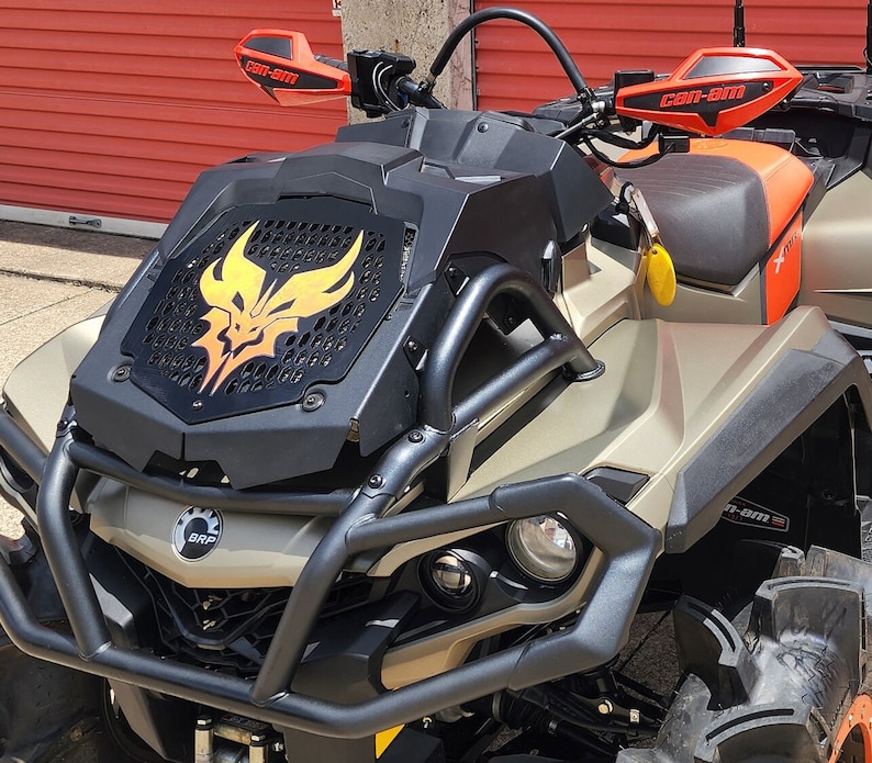 May include: A tan and black Can-Am off-road vehicle with a black and yellow graphic on the front grill. The vehicle has a roll cage and a black seat. The vehicle is on a black surface.