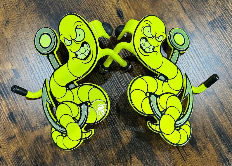 K&ouml;nnte beinhalten: Zwei neon-gelbe Wandhaken aus Metall mit einem Cartoon-Wurm mit w&uuml;tendem Gesichtsausdruck, der sich um einen Angelhaken windet. Jeder Haken hat ein geschwungenes Design mit schwarzen Gummispitzen an den Enden und einer runden Montageplatte.