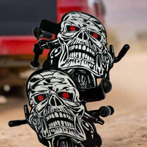 K&ouml;nnte beinhalten: Zwei schwarze Skateboard-Achsen aus Metall mit einer Grafik eines wei&szlig;en Totenkopfes mit roten Augen. Das Totenkopf-Design weist schwarze Liniendetails auf und befindet sich auf der Vorderseite jeder Achse. Die Metallteile sind schwarz gl&auml;nzend.