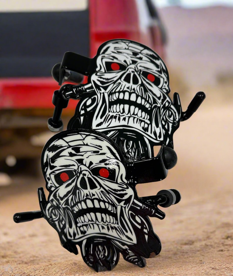 K&ouml;nnte beinhalten: Zwei schwarze Skateboard-Achsen aus Metall mit einer Grafik eines wei&szlig;en Totenkopfes mit roten Augen. Das Totenkopf-Design weist schwarze Liniendetails auf und befindet sich auf der Vorderseite jeder Achse. Die Metallteile sind schwarz gl&auml;nzend.