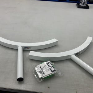 May include: Two white, curved metal support arms with cylindrical stems and a small, clear plastic-wrapped package of green Rip-Tie cable wraps positioned between them. Each metal arm features a small rectangular label on the curved section.