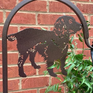 May include: A metal silhouette of a brown dog with a curly tail, mounted on a metal arch. The silhouette is against a brick wall with green plants in the background.