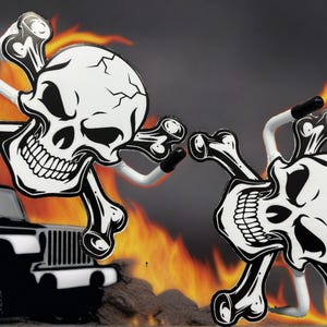 K&ouml;nnte beinhalten: Zwei Wandhaken aus Metall in Form von Totenk&ouml;pfen mit gekreuzten Knochen in Schwarz und Wei&szlig; mit Rissmuster. Jeder Haken verf&uuml;gt &uuml;ber einen wei&szlig;en Metallarm mit schwarzer Gummispitze zur Montage, vor einem Hintergrund aus Flammen und einem schwarzen Fahrzeug.
