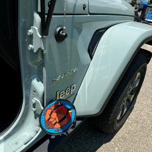 K&ouml;nnte beinhalten: Ein hellgraues Jeep Wrangler Sahara Fahrzeug mit einem kreisf&ouml;rmigen Grafikaufkleber am unteren T&uuml;rscharnier. Der Aufkleber zeigt eine rote Teufelsfigur mit H&ouml;rnern und Schwanz vor einem blauen Hintergrund mit schwarzem Netzmuster.