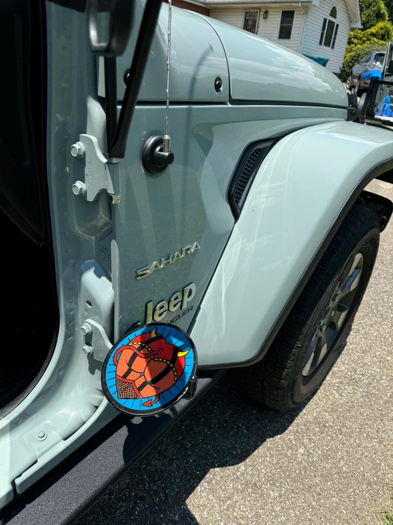 K&ouml;nnte beinhalten: Ein hellgraues Jeep Wrangler Sahara Fahrzeug mit einem kreisf&ouml;rmigen Grafikaufkleber am unteren T&uuml;rscharnier. Der Aufkleber zeigt eine rote Teufelsfigur mit H&ouml;rnern und Schwanz vor einem blauen Hintergrund mit schwarzem Netzmuster.