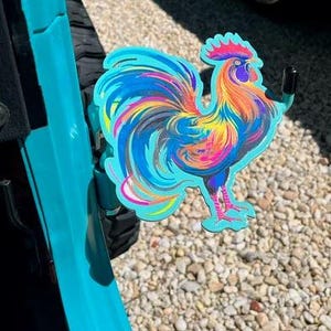 K&ouml;nnte beinhalten: Ein leuchtend buntes, hahnf&ouml;rmiges T&uuml;rscharnier-Zubeh&ouml;r aus Metall mit einem Farbverlauf in T&uuml;rkis, Gelb, Orange und Magenta. Die Hahnfigur ist an einem schwarzen Metallhaken befestigt, der an einem t&uuml;rkisfarbenen Fahrzeugt&uuml;rscharnier montiert ist.