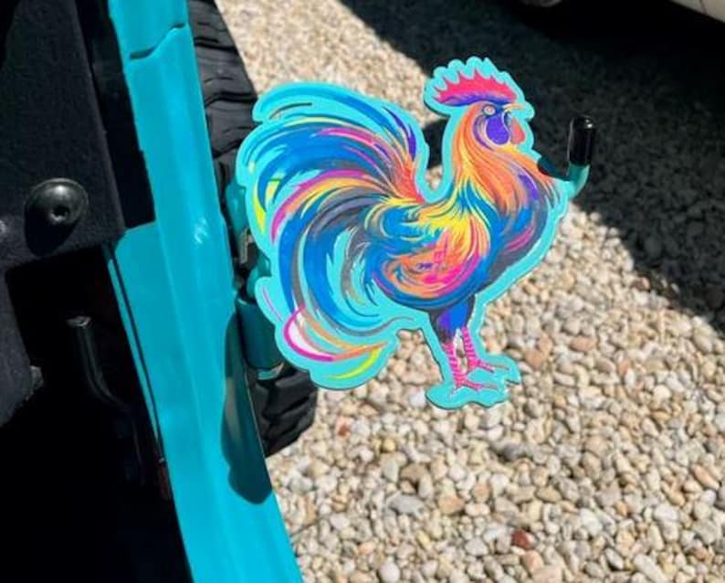 K&ouml;nnte beinhalten: Ein leuchtend buntes, hahnf&ouml;rmiges T&uuml;rscharnier-Zubeh&ouml;r aus Metall mit einem Farbverlauf in T&uuml;rkis, Gelb, Orange und Magenta. Die Hahnfigur ist an einem schwarzen Metallhaken befestigt, der an einem t&uuml;rkisfarbenen Fahrzeugt&uuml;rscharnier montiert ist.