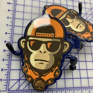 K&ouml;nnte beinhalten: Zwei Wandhaken aus Metall mit einer gl&auml;nzenden Grafik eines Affen, der eine orangefarbene Baseballkappe, eine dunkle Sonnenbrille und eine Kette mit einem runden Anh&auml;nger tr&auml;gt, platziert auf einer Schneidematte mit Zentimeter-Skala.