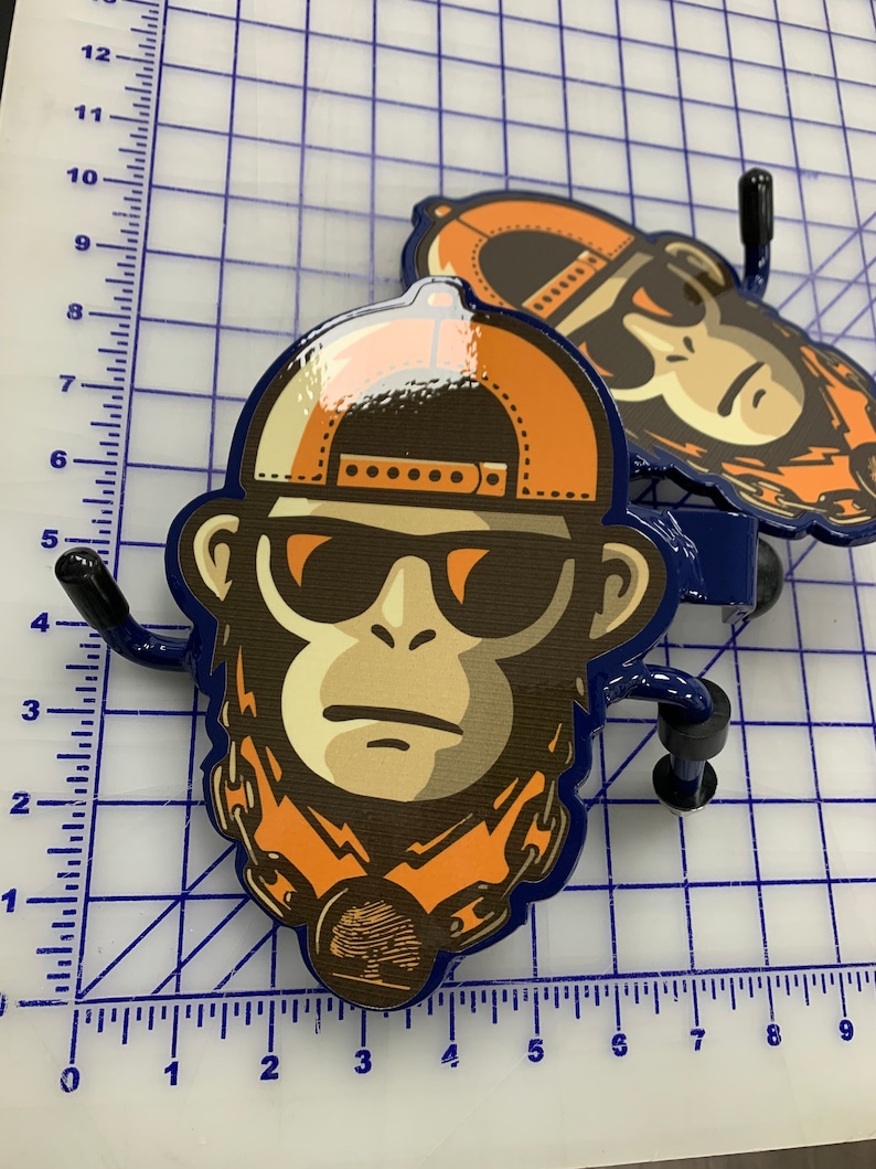 K&ouml;nnte beinhalten: Zwei Wandhaken aus Metall mit einer gl&auml;nzenden Grafik eines Affen, der eine orangefarbene Baseballkappe, eine dunkle Sonnenbrille und eine Kette mit einem runden Anh&auml;nger tr&auml;gt, platziert auf einer Schneidematte mit Zentimeter-Skala.