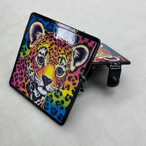 K&ouml;nnte beinhalten: Zwei quadratische Anh&auml;ngerkupplungsabdeckungen aus Metall mit einer lebendigen, farbenfrohen Illustration eines Leopardenjungen-Gesichts. Das Design umfasst helle Gelb-, Orange-, Rosa- und Blaut&ouml;ne mit schwarzen Flecken. Jede Abdeckung hat einen schwarzen Rahmen.