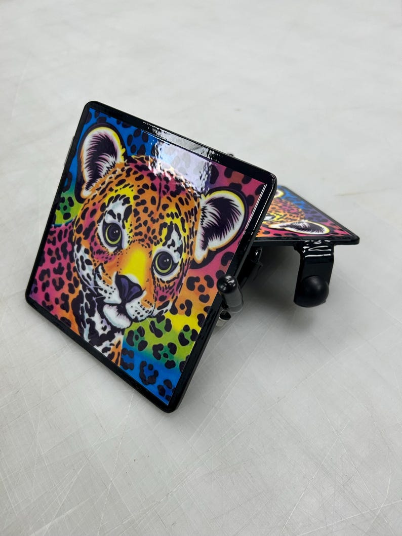 K&ouml;nnte beinhalten: Zwei quadratische Anh&auml;ngerkupplungsabdeckungen aus Metall mit einer lebendigen, farbenfrohen Illustration eines Leopardenjungen-Gesichts. Das Design umfasst helle Gelb-, Orange-, Rosa- und Blaut&ouml;ne mit schwarzen Flecken. Jede Abdeckung hat einen schwarzen Rahmen.