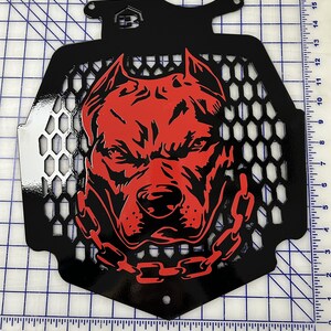 K&ouml;nnte beinhalten: Schwarzer Metallgrill mit einem roten Pitbull-Hundegesichtsdesign. Der Grill hat ein Wabenmuster und eine Kette um den Hals des Hundes.