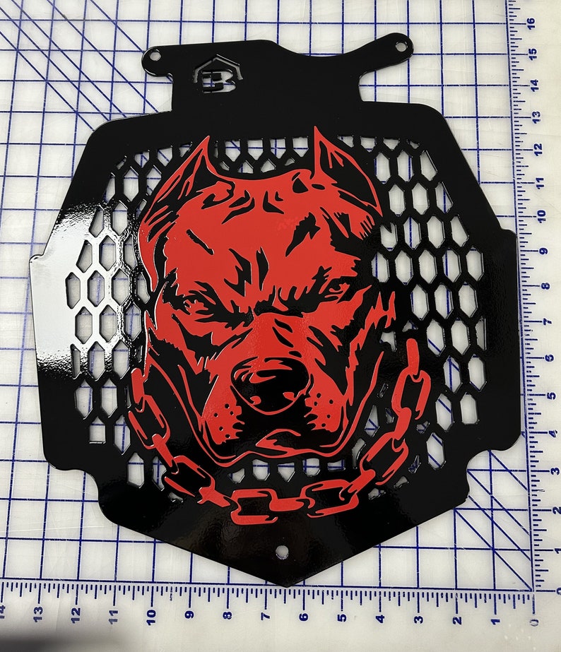 K&ouml;nnte beinhalten: Schwarzer Metallgrill mit einem roten Pitbull-Hundegesichtsdesign. Der Grill hat ein Wabenmuster und eine Kette um den Hals des Hundes.