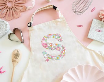 Puede incluir: Un delantal de lona blanco con una letra "S" estampada con un patrón floral en la parte delantera. El delantal tiene correas ajustables y un bolsillo.