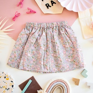 Puede incluir: Falda con estampado floral y cintura elástica. La falda presenta un patrón de pequeñas flores rosas y azules sobre un fondo rosa claro. La falda está fruncida en la cintura y tiene forma acampanada. La falda es probablemente para una niña.