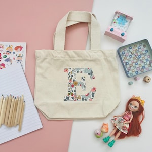 May include: A cream tote bag featuring a floral "E" design, with a doll, toy games, and pencils. The bag has a sturdy handle and is made of a canvas-like material. The background is a mix of pink and white.