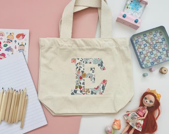May include: A cream-colored tote bag with a floral "E" design, alongside a doll, toy games, and colored pencils. The bag has a sturdy handle and is made of a canvas-like material. The background is a mix of pink and white.