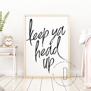 Puede incluir: Una impresión de arte de pared imprimible en blanco y negro con el texto "keep ya head up" en estilo manuscrito. La impresión está enmarcada en un marco de madera claro y se muestra en una pared blanca.